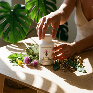Hand reaching for the Liver+ bottle on a table with milk thistle, dandelion, and artichoke