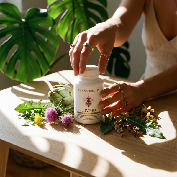 Hand reaching for the Liver+ bottle on a table with milk thistle, dandelion, and artichoke